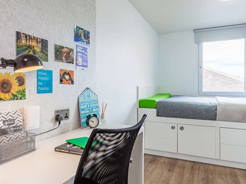 A well-lit student room with a bed, study area, and natural light coming through the window.